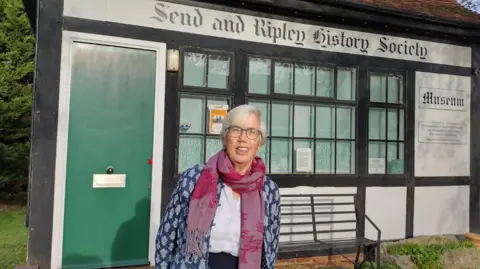 Fiona McCarthy A woman with short grey hair and glasses wearing a purple scarf and a blue and white cardigan. She is standing outside a building with a green door, the sign above which says: Send and Ripley History Society.
