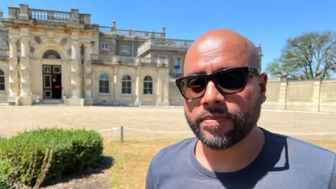 Mousumi Bakshi/BBC A man wearing sunglasses and a T-shirt while standing in front of a grand country house.