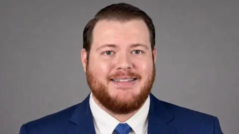 Yeovil Conservatives A man with short, coiffed hair, and a short ginger beard is smiling at the camera. He is wearing a blue suit and tie. 