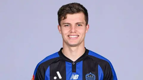 Young footballer Callan Elliot with dark hair smiles in the blue and black striped kit of Auckland FC