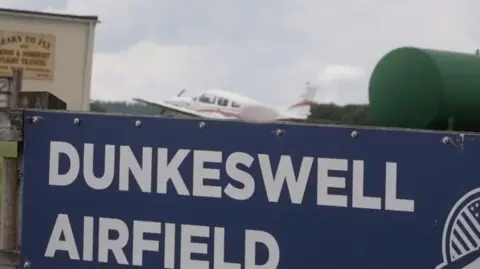 A picture of Dunkeswell Airfield sign. It is blue and white in colour, there is a white plane in the distance.