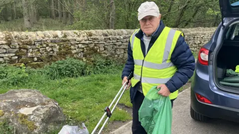 BBC/Grace Wood A man in a grey baseball cap and yellow high vis jacket picks up litter from a verge