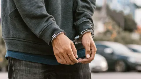 Under arrest, a man's hands with clenched fists are handcuffed behind him. 