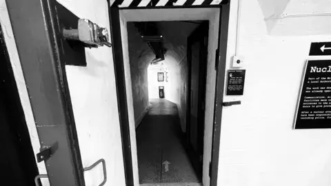 Black and white photo of a large open door made of thick reinforced metal with a large lock. Beyond the door is a long curved corridor or tunnel.