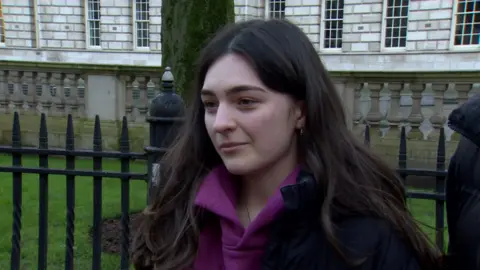 A young woman with long brown hair is wearing a purple hoody and black coat. She is standing in front of a set of black railings and a white bricked building.