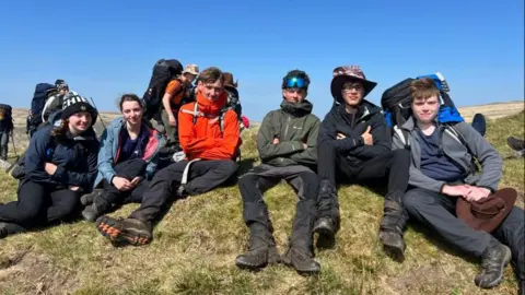 L-R Abbie, Jess, Aiden, Roody with other participants sat on the grass together with their rucksacks on their back.