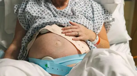 A pregnant woman lies in a hospital bed in the UK with monitors strapped to her stomach. She is propped up with a pillow and is wearing a hospital gown with white sheets draped over her legs