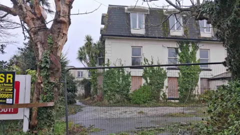 States of Guernsey The outside of a derelict hotel in Guernsey which is boarded up and closed off and is completely overgrown by greenery. 