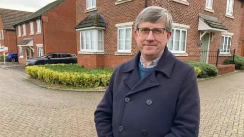 A man in a blue coat and glasses is standing outside a house on a new build estate