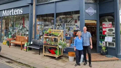 David and Sharron Jowett stand arm in arm outside a shop front. The shop front is navy and has plants and gardening supplies on a wooden stall outside. Hardware products can be seen in the front window. The name 'Mortens' is above the shop door.