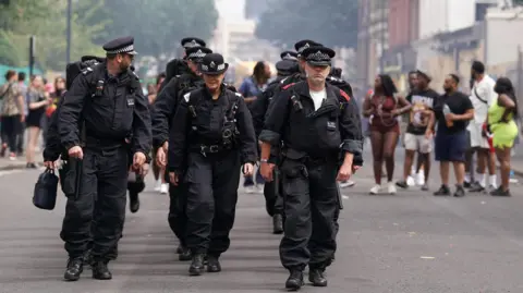 PA Media A group of police walk three abreast past groups of revellers along a London street. The police are wearing black riot boiler suits. 