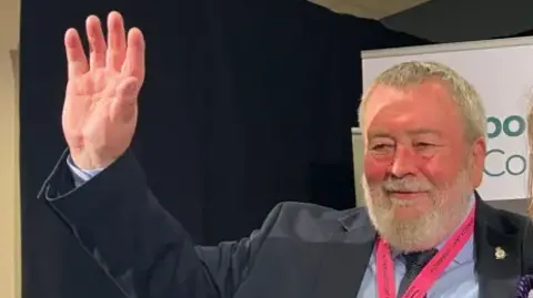 Shariqua Ahmed/BBC John Fox wearing a dark grey suit and a blue shirt and a bright pink lanyard around his neck- waving is hand