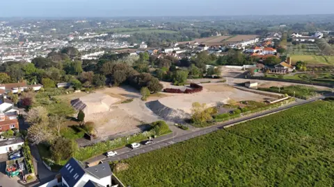 BBC Drone shot overlooking the Overdale Acute Hospital development site in St Helier. Large mounds of building material are at the site. The area is surrounded by trees, fields and buildings.