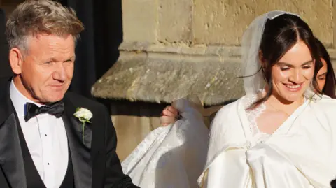 PA Media Gordon Ramsay pictured entering Bath Abbey with his daughter Holly. He is wearing a black suit and she is wearing a white dress and veil. 