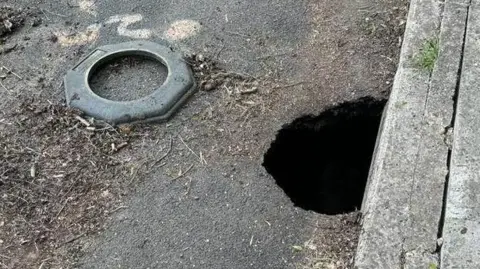 Steve Baker A road in Castle Cary, Somerset, with a hole on the side of the road right next to the pavement.