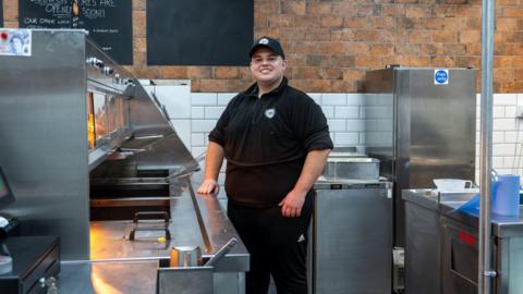 Stephanos Constantinou, manager of The Island House Fish and Chip Shop standing beside the metal food counters in black uniform