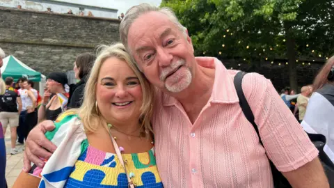 A woman wearing a colourful top smiles at the camera. Next to her is a man in a short-sleeved pink shirt. He has his right arm around the woman's shoulder. 