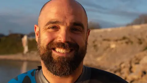 Jason with dark hair and beard, wearing a blue and black swimming wetsuit