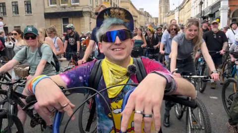 Wood is wearing ski-like goggles and a trippy Simpsons t-shirt. He has facial piercings and lots of rings.