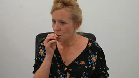 A woman with auburn hair in a top bun hair style tastes a piece of chocolate. She is sitting on a chair and is wearing a black dress, which has blue and orange flowers on it. 