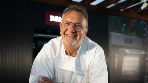 Getty Images Raymond Blanc, wearing his chef's whites and glasses, smiles at the camera. He has short grey hair and a short beard, and his arms are folded across his chest