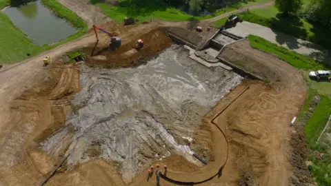 Splice Films An aerial view of a pond, showing mud, and people standing in it. There is greenery around it and diggers. Work is being carried out and the pond is empty of water. 