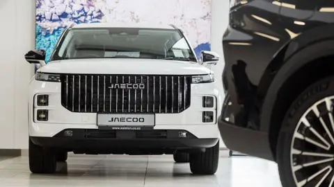 Getty Images A white SUV sits on the floor of a car sales showroom, with the rear of a black SUV sitting in the foreground.