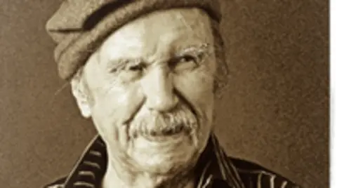 Ulster History Circle A black and white phot of Padraic Fiacc. He has a moustache and is wearing a woollen cap and a striped shirt.