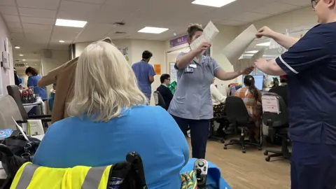 A lady say in a wheelchair with hospital workers passing documents between them and others working on computers in the background.