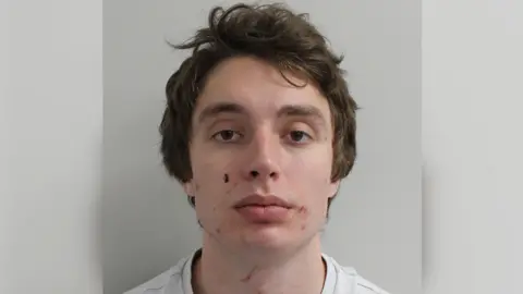 Custody image of Matvei Rumiantsev. He is a young man with short brown hair, wearing a white t-shirt. He stands with a neutral expression in front of a white wall.