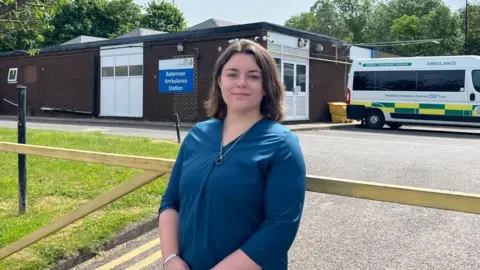 Sophie Crossthorn A woman with dark brown hair in a shoulder length bob is wearing a blue top and silver necklace. She is standing outside an ambulance station with a building and an ambulance in the background
