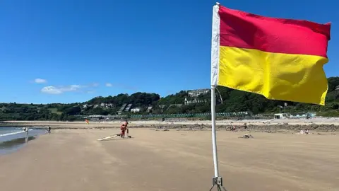 A red and yellow beach flag indicating one end of a lifeguard monitored bathing zone on Langland beach flies in the foreground of the picture. In the near distance a lifeguard sits on a high chair supervising the water and talking to someone with a surfboard. A wide stretch of sand and some rocks is visible as the tide is at its lowest point. In the far distance the row of white and green beach huts can be seen above the edge of the beach. A building housing a cafe is visible just under the flag, while above the beach, trees and green fields can be seen, with some flats and houses interspersed among the greenery. The sky is light to deeper blue with a few wispy clouds sitting low over the horizon.