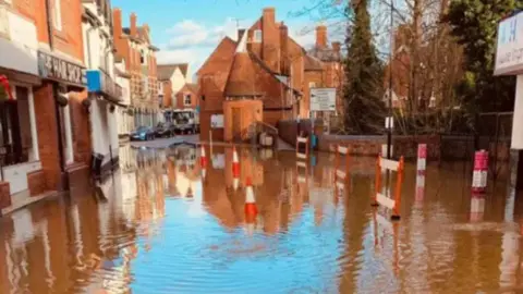 Worcestershire County Council A flooded street in a town centre, with orange and white traffic cones and barriers placed in the waters. Redbrick buildings line the streets.
