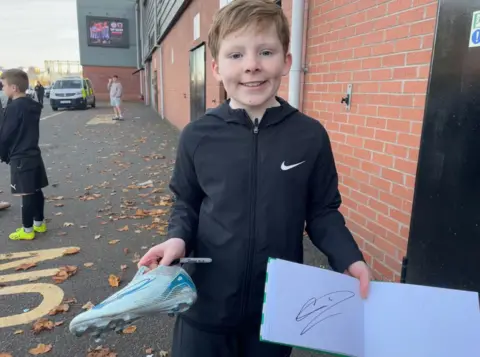 Zac wears a black Nike tracksuit and holds a signed boot and notebook outside of the St Mirren FC stadium 