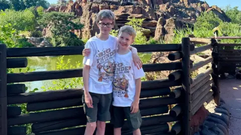 Supplied Two boys aged 10 and thirteen with their arms around each others shoulders are posing for the camera. They are wearing white patterned t-shirts and dark green shorts. The older child is on the left and is wearing glasses.