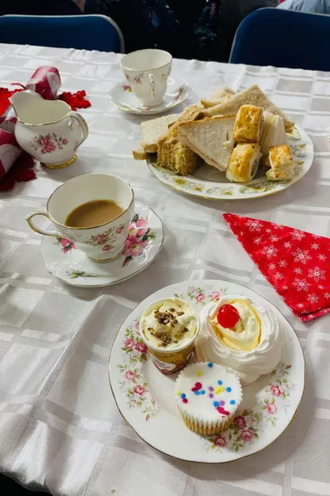 SARAH CALLOW An spread for afternoon tea laid out on delicate china plates and cups. There are three cream cakes, a cup of tea, a milk jug and a plate of sandwiches and pastries on a white tablecloth that also has a red festive paper napkin on it.