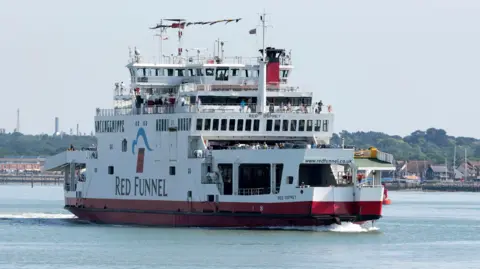 Getty Images A large white ferry with "RED FUNNEL" printed on the side is pictured in motion as it crosses a body of water. People can be seen on the deck looking out from the railings. 