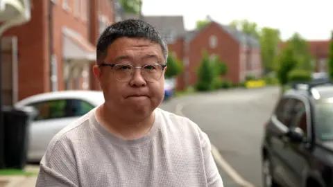 Jonathan Yu, a man wearing glasses, is stood on a driveway with a housing estate behind him. There is a black car behind him to one side, and a white car to the other side which is side-on and appears to be parked on a driveway.