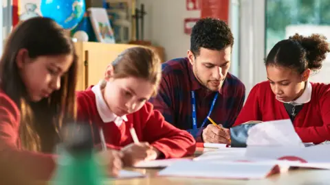 Getty Images Three school girls and a teacher sat at a table. Two girls are sat to the left of a teacher and one is to the right. The girls are wearing red tops while the teacher is in a red and blue shirt. He has a lanyard over him that says staff in capital letters. The girls are writing in note pads while the teacher helps the girl to the right.