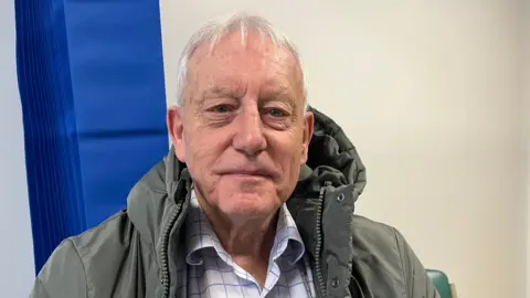 Ellen Knight/BBC Kendall photographed looking into the camera and smiling slightly. He is wearing a white collared shirt with blue checks, and a hooded khaki coat. He has short silver hair. He is sat down in a room with cream walls, and behind him is a royal blue privacy curtain. 