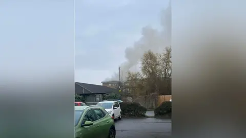 Lucas A picture of a residential area and in the background grey smoke is being emitted from a block of flats