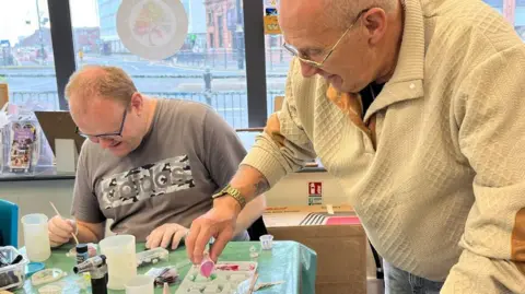ArtWorks Art4All Two men work on an art project at one of the Men's Make and Mend sessions. The man on the left is sitting down and appears to be using a thin brish of some type. He has short fair hair and is wearing glasses and a grey Adidas T-shirt. The man on the right is standing and also has glasses. He is wearing a beige top. He is pouring paint from a small tub.