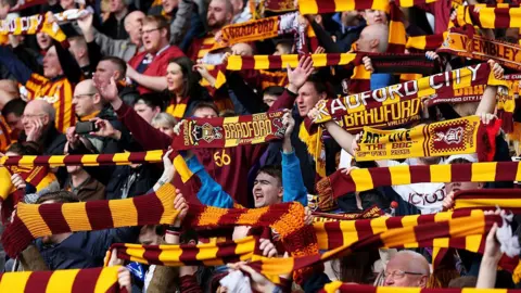 A crowd of Bradford City football fans holding claret and amber striped scarves in the air.