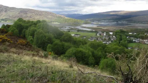 The Highland Council The image is taken from a hillside above Fort William. Loch Linnhe and rolling hills are visible in the distance. Nearby there are gorse bushes and trees on the slope. 