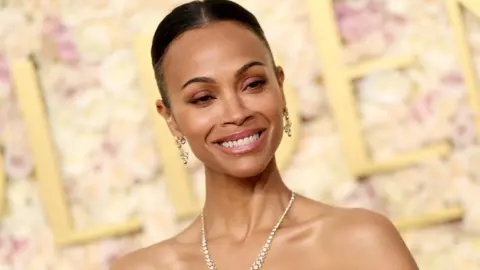 Zoe Saldaña attends the 82nd Annual Golden Globe Awards at The Beverly Hilton on January 05, 2025 in Beverly Hills, California. She is smiling and is wearing a diamond necklace and matching drop earrings. 