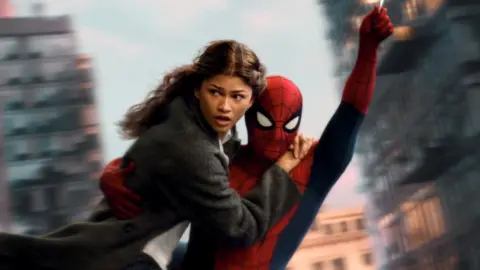 Spider-Man swings through the city on web whilst holding MJ, played by Zendaya.
