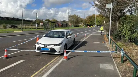 Ben Mellor/BBC A white taxi with police tape around it. A lone police officer is stood in the background.
