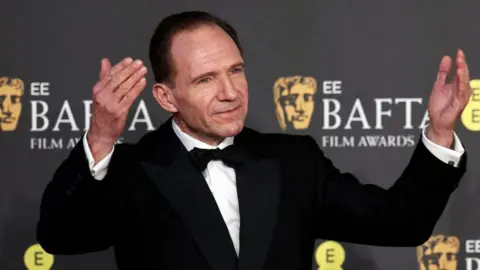 Ralph Fiennes stood with his hands raised to either side of his head dressed in a tuxedo