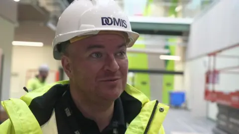 A man wearing a white hard hat and hi-vis jacket, speaks and looks off camera, in a youth zone which is being built