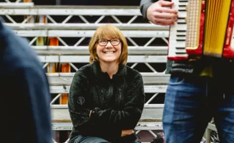A woman wearing a dark-coloured jumper is sitting down and smiling while watching a man playing an accordion. There is a metal structure behind the woman.